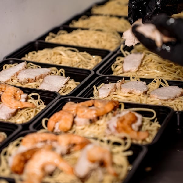 A gloved hand adds chicken to a row of Victoria's Kitchen & Catering meal prep containers with pasta and shrimp.
