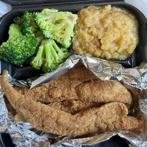 Fried fish, steamed broccoli, and mashed potatoes in a black takeout container from Victoria's Kitchen.