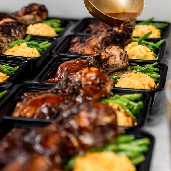 A ladle pours sauce over Victoria's Kitchen's soul food meal prep containers with chicken, green beans, and mashed potatoes.