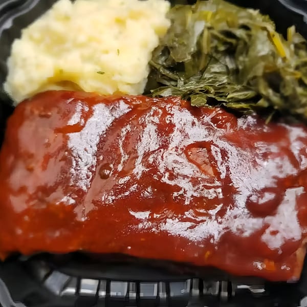 A soul food plate with BBQ ribs, mashed potatoes, and collard greens in a black container.