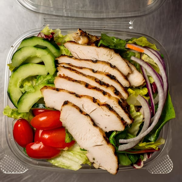 Grilled chicken salad with cucumbers, tomatoes, red onion, and mixed greens in a plastic container.